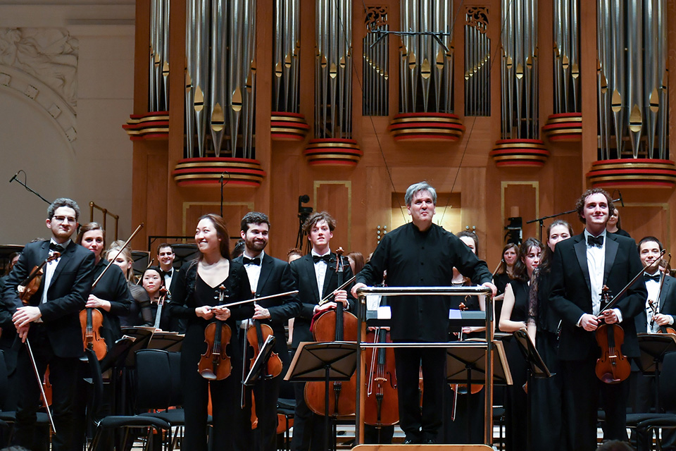 Sir Antonio Pappano, a man wearing formal attire, smiling facing the audience with the students in the orchestra, holding their instruments, in the RCM's Amaryllis Fleming Concert Hall